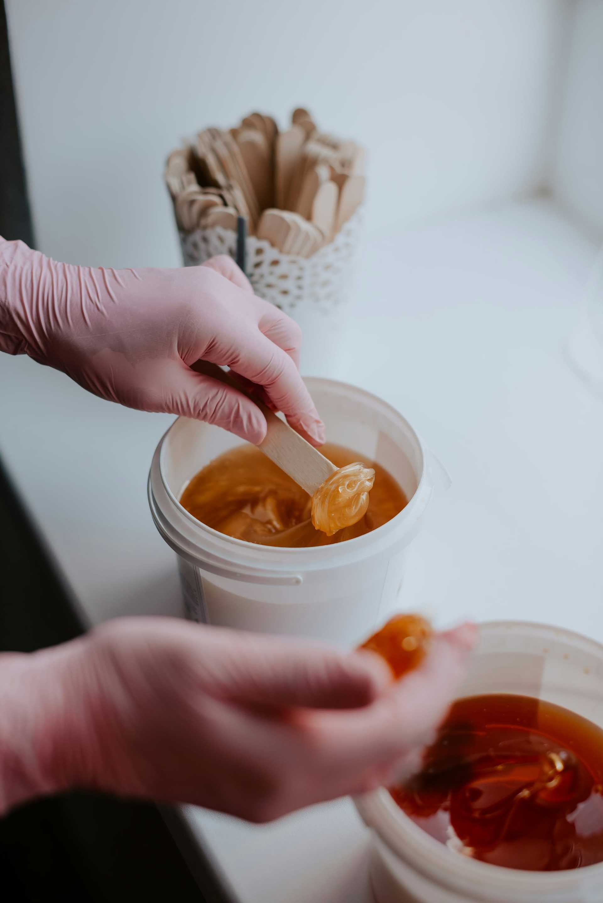 Hands in gloves preparing sugar wax with wooden sticks.